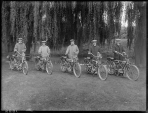 Five unidentified men in riding attire standing with Singer motorbikes on stands, on grass under large willow trees with park bench [Hagley Park?] and [the Avon River?] behind, Christchurch