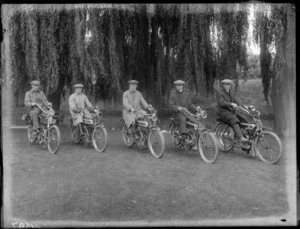 Five unidentified men in riding attire sitting on Singer motorbikes on stands, on grass under large willow trees with park bench [Hagley Park?] and [the Avon River?] behind, Christchurch