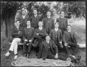 Group of unidentified men dressed in business attire, [members of a tug o' war team?], with a coiled rope at their feet, in a park, probably Christchurch district