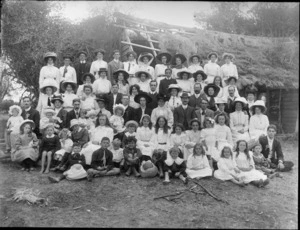 Large group of unidentified people, including a number of children, sitting outside a building with a partially thatched roof, probably Christchurch district
