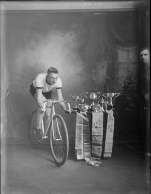 Studio portrait of unidentified cyclist on a bicycle, with a table of trophies and ribbons beside him, showing unidentified man on the right side, taken in the studio, probably Christchurch district