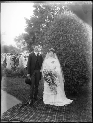 Wedding portrait, showing an unidentified bride and groom, in an outdoor location, with members of wedding party visible in distance, possibly Christchurch district