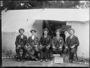 Group of five young men at a campsite, sitting alongside tents, with sign reading 'Myrtle Camp' behind, and a sign reading 'Please Keep Off The Grass' in front, possibly Christchurch district