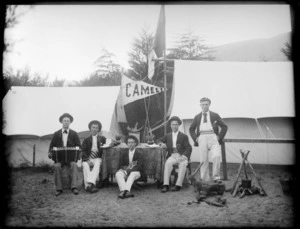 Group of unidentified young men at a campsite, one with an accordian, another with a kitten, and two others reading books sitting at a table outddors, a pennant reading 'Camelia' hanging between tents behind, possibly Christchurch district