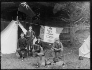 Four young men, one with an accordian, sitting by a campfire, next to two tents, including a sign with the words 'Ivy Camp' in decorative script, possibly Christchurch district