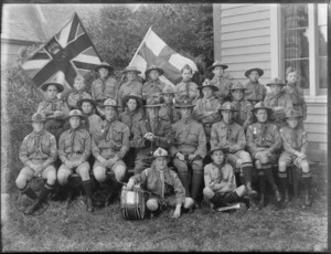 Unidentified Boy Scout troop in uniform, in the backyard in front of wooden house, with twenty young boys, four older boys and one adult male, with two flags, a bugle and drum, probably Christchurch region