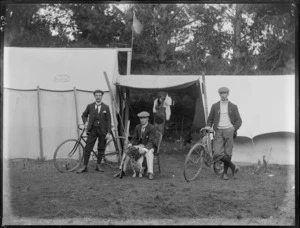 Group of three unidentified men at a campsite, two with bicycles and one with a dog and gun, alongside tents, one marked with a stamp reading 'J Trist, Maker, Christchurch', possibly Christchurch district