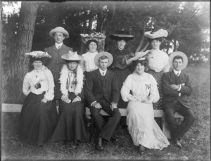 A group of unidentified people, six women and three men, showing three women and two men sitting on a wooden bench, three women and a man standing at the back under the trees, possibly Christchurch district