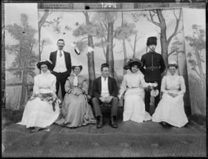 Unidentified actors in front of false backdrop, on grass with brick wall behind, man with false moustache and young lady, older couple, young woman with bonnet and umbrella, maid, soldier with fur hat and false moustache, probably Christchurch region