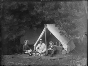 Three unidentified men sitting in front of the tent having a cup of tea, showing a wooden chest, an accordion and a long handle pan, in a unidentified camping ground, possibly Christchurch district