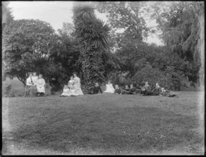 A group of unidentified women and children, showing some of the children lying down on the grass, some of the children sitting down on the grass, and the two women sitting on chairs with some of the children, in the garden at the back of the house, possibly Christchurch district