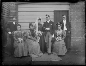 Wedding portrait of unidentified bride and groom with wedding party, showing three of the women sitting, at the back of the house, possibly Christchurch district