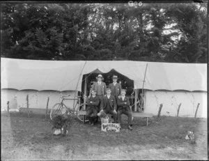 Five unidentified members of the Imperial Camp, with three men sitting on a wooden bench, with a dog in front of them and two men standing behind them in front of a tent, showing a sign for Imperial Camp and a bicycle in the background, probably Christchurch district