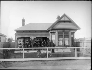View of the front porch of the house from an unidentified street, probably Christchurch district, showing nine women and six men on the porch, with two bicycles against the fence on the right side and a sign Te Aro, above the front windows
