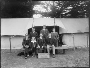 Six unidentiified members of the Imperial Camp, three men sitting on a wooden bench, with a boy in front of them and a dog asleep under the bench on the left side, three men standing behind them in front of an open tent, possibly Christchurch district