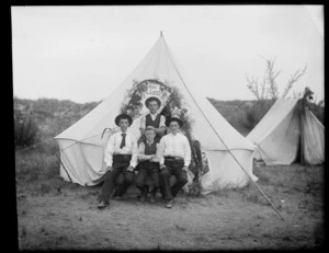 Four unidentified members of the Huia Camp, possibly Christchurch district, showing three boys sitting on a wooden bench, with one boy standing behind them in front of the tent, with a fern decoration and the sign for Huia Camp on the front flap of the tent