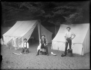 Three unidentified men at a campsite in front of two tents doing odd jobs, showing one man chopping wood, one man doing dishes and the other man cooking, possibly Christchurch district