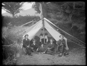 Five unidentified men in front of an open tent, showing a table with a floral arrangement and cards on it and wooden benches, possibly Christchurch district, showing one man sitting on a wooden stool playing an accordion, one man lying down in the front with two men sitting behind him on the ground and the other man sitting on a wooden crate