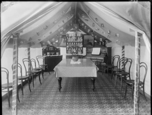 Interior view of a tent, showing a table, chairs, a piano, a china cabinet with cups, plates and bowls, a sideboard dresser with jugs and bowls and prints on the back wall, possibly Christchurch district