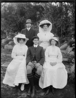 Five unidentified people, three women and two men, showing a man and two women sitting on a wooden bench and one man and one woman standing behind them, in a unidentified park, possibly Christchurch district