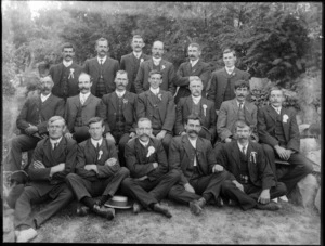 A group of unidentified men, showing five men sitting on the grass in the front, seven men sitting on a wooden bench and six men standing behind them, in an unidentified park, possibly Christchurch district