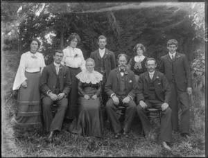 Members of a unidentified family, showing five men and four women, with three men and a woman sitting on chairs and three women and two men standing behind them, in front of a macrocarpa, possibly Christchurch district
