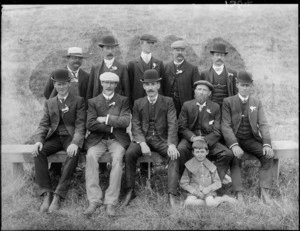 A group of unidentified men and a boy, showing the boy sitting on the grass in the front, five men sitting on a wooden bench and five men standing behind them, in an unidentified park, possibly Christchurch district