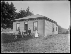 Unidentified family group in front of the house, showing a woman sitting on the front step, a man standing with a bicycle, a girl standing with a bicycle, a man sitting on the grass with a dog, a boy standing with a bicycle, a boy in a pushchair and a girl with a doll's pram, possibly Christchurch district