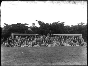 A group of unidentified people, showing men, women and children sitting and standing in front of two tents, in an unidentified park, possibly Christchurch district