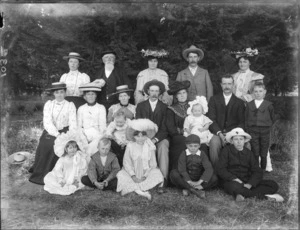 Unidentified family portrait, four men, seven women, four boys, two girls and two babies, showing the children sitting on the grass in the front, two men and four women sitting on a wooden bench, two of the women holding the babies, three women and two men standing behind them, possibly Christchurch district