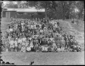 A group of unidentified men, women and children, sitting on a bank below a shed in a unidentified park, possibly Christchurch district