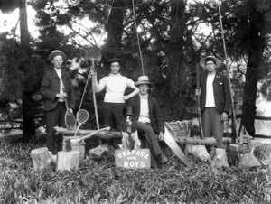 Four young men and a dog, with a variety of equipment including a gun, an axe and a sign saying 'Okapara Boys'