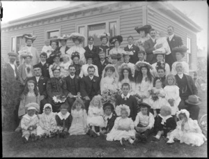 Wedding portrait of unidentified bride and groom with wedding party and family members in the garden beside a house, possibly Christchurch district