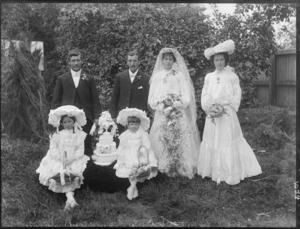 Unidentified wedding group outdoors, showing bride and groom, wedding party and three-tier wedding cake, probably Christchurch district