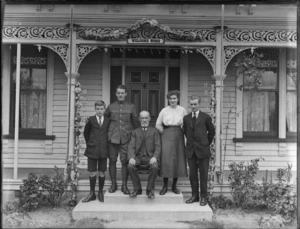 Unidentified family group, including a man in an army uniform on porch decorated with small Union Jack flags, includes 'Welcome Home' sign, probably Christchurch district