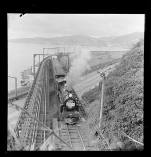 A steam train on a rail bridge over Wellington Motorway, on the occaision of the 50th anniversary of the Manawatu Railway, including Lambton Harbour in background