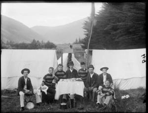 Group of unidentified men dining outside a tent, probably Sumner, Christchurch, sign above them says 'Rovers Camp', includes a piano accordian in the front with a kettle on top