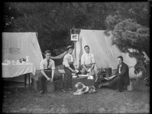 Group of unidentified young men preparing food outside tents, J Welsh, Maker, Christchurch is written on one tent with a dog lying in front on grass and a framed photograph hangs from another, probably Christchurch district