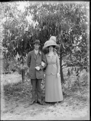 Full portrait of unidentified man and woman, standing beneath gum trees, probably Christchurch district, woman wears a large hat with ostrich plumes