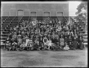 Large group portrait of unidentified people with hats on grass and on bleachers (seats) with tall brick building behind [sports ground?], probably Christchurch region