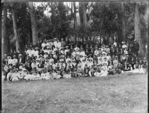 Very large group portrait of unidentified people on social outing under gum trees, with hats and brass band instruments and children in front, probably Christchurch region