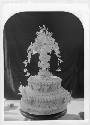 Studio portrait of two layered wedding cake with intricate icing patterns and basket of flowers on top, Christchurch