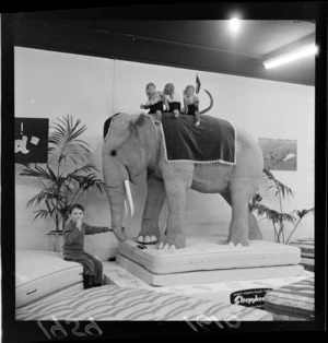 An unidentified small boy sitting alongside a large stuffed elephant and monkeys on top of a sleephead mattress at the Wellington Industries Fair exhibits
