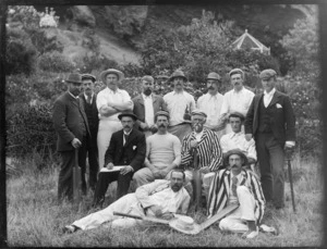 Group portrait of fourteen unidentified men with hats and cricket bats, on long grass with low hedge and building with garden behind and steep hill above, [Sumner?], Christchurch