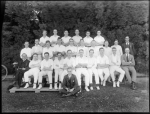 Large group portrait of unidentified cricket team, players in whites and coaches, with one man in front holding a cricket ball, on grass in front of tall trees, probably Christchurch region