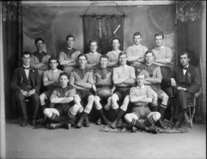 Studio portrait of the Woolston junior men's rugby league football team for 1919, unidentified members and coaches with rosettes, with ball in front and club banner hanging behind, Christchurch
