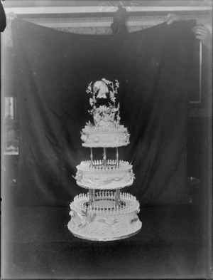 Three layered wedding cake with pillars, intricate icing patterns and flowers arrangement with bow on top, with false backdrop, probably Christchurch region