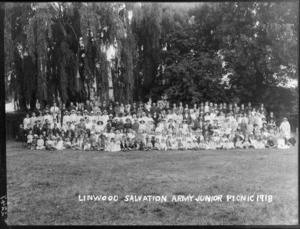 Very large group portrait of unidentified men and women in hats, children in front, with men in military uniforms, on grass under tall trees, picture inscribed with 'Linwood Salvation Army junior picnic 1918', Christchurch