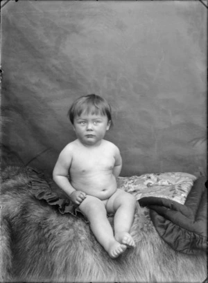 Studio portrait of unidentified naked baby sitting on fur rug and cushion, Christchurch