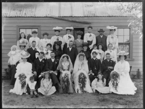 Large double wedding unidentified group portrait, two brides with long veils and grooms with extended families, children and babies, in front of wooden building with 'Hinemoa' sign, probably Christchurch region